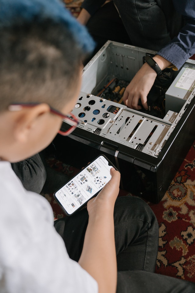 A man looking into his phone while another man is fitting components inside a PC. Concept image of buying PC upgrades. Concept image to show weight of gaming PC.