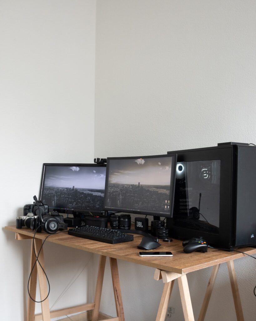 A wooden table having 2 black colored monitors and a gaming PC with some keyboars, mice and console controllers.